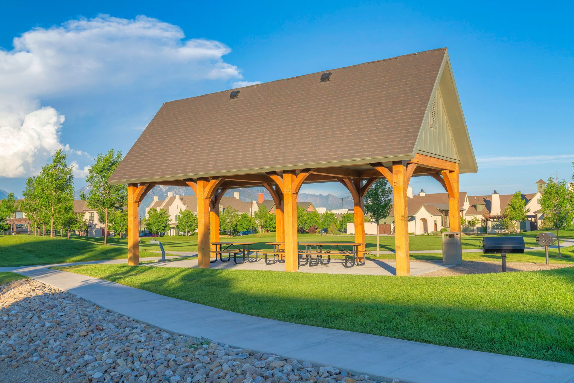 Wooden pavilion with picnic tables on green grass under a blue sky in a park.