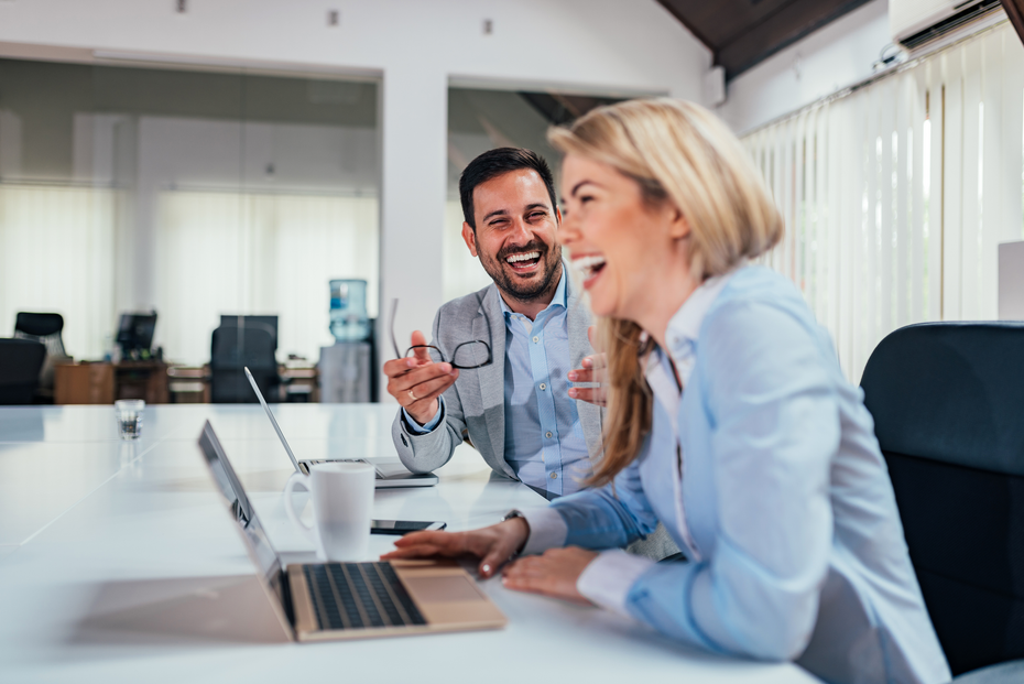 A man and a woman laughing at something on a laptop in an office setting.