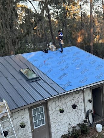 Person on a roof installing a blue-covered metal roof; house exterior with ladder in the background.
