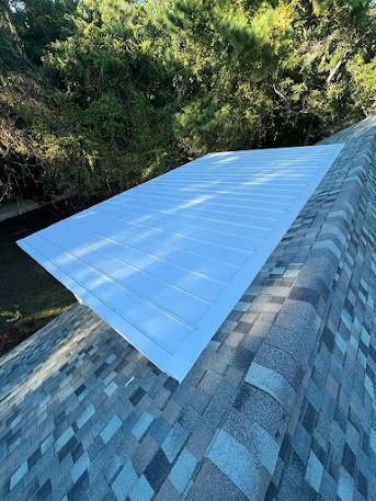 A white skylight on a shingled roof, viewed from an angle, with trees in the background.