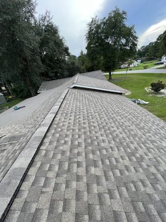 Gray shingled roof on a house, surrounded by green trees and grass under a cloudy sky.