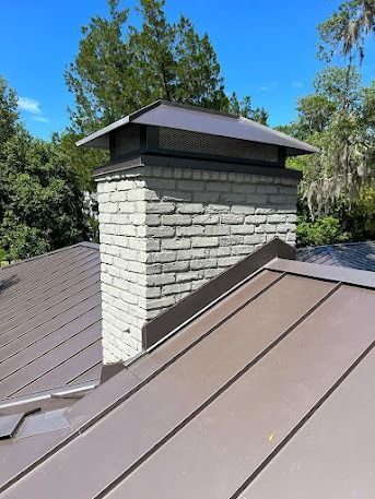 Chimney with stone veneer and metal cap on a brown metal roof under a blue sky.