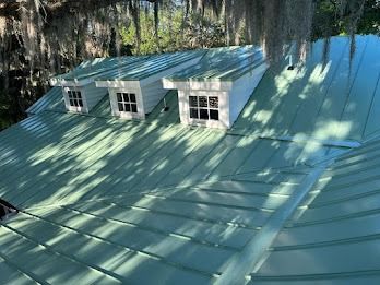 Green metal roof with three dormer windows in a wooded setting.