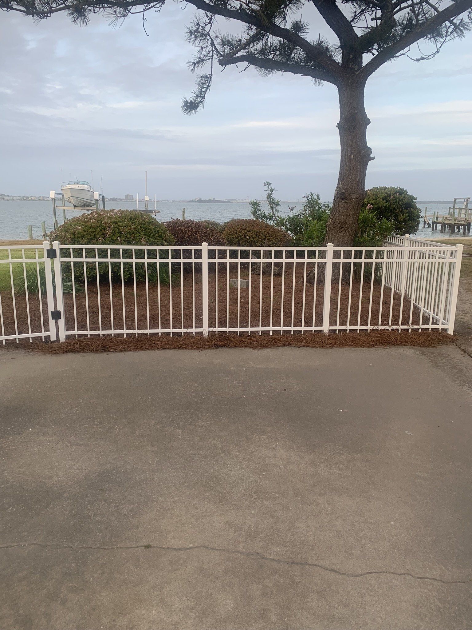 White fence in front of bushes and a tree, with a body of water and boats in the background on a cloudy day.