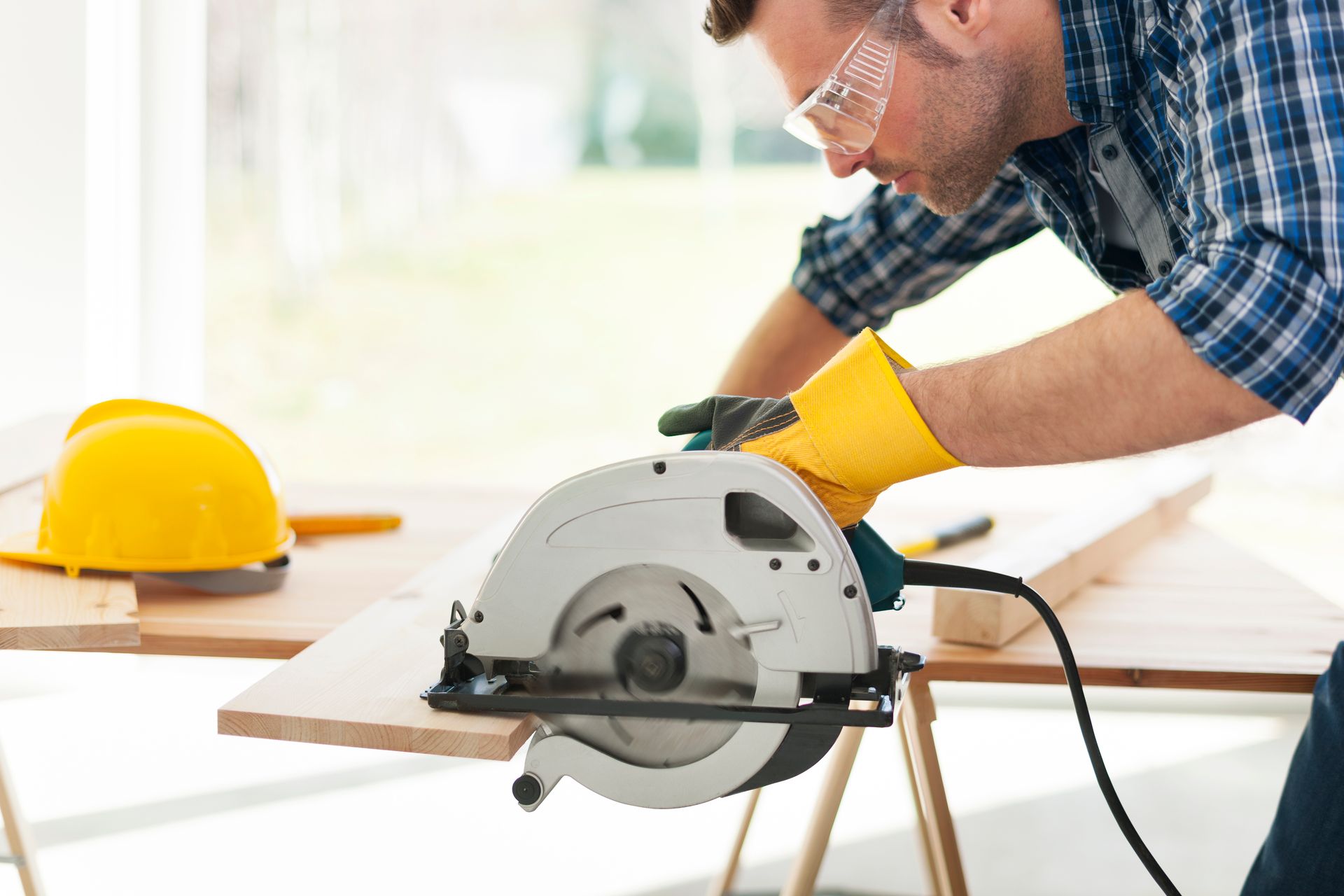 Worker cutting wooden board with circular saw, safety gloves and helmet on table.