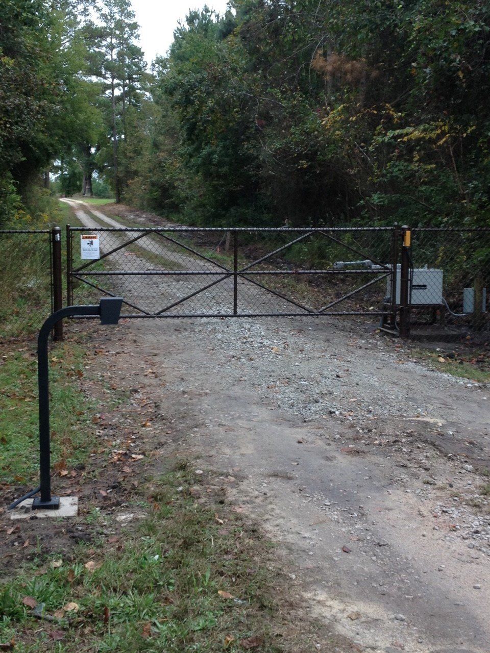 Black metal gate across a gravel road leading into a wooded area. An automated gate system is visible.
