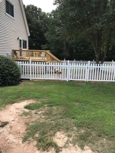 White picket fence surrounds a backyard with a deck, dog visible near gate, beside a beige house.