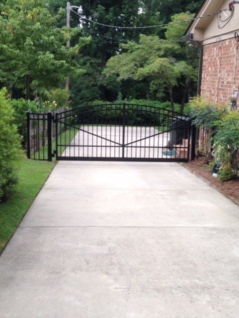 Black wrought iron gate on a concrete driveway, leading to a house with brick facade and lush greenery.