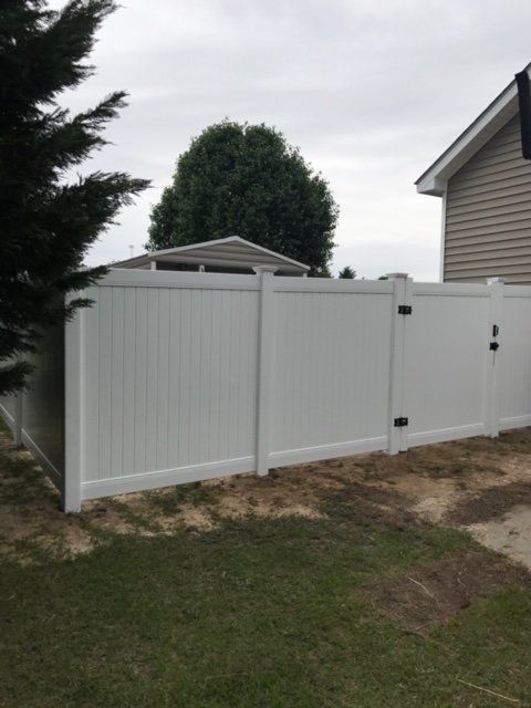 White vinyl fence with gate surrounding a yard next to a house under a cloudy sky.
