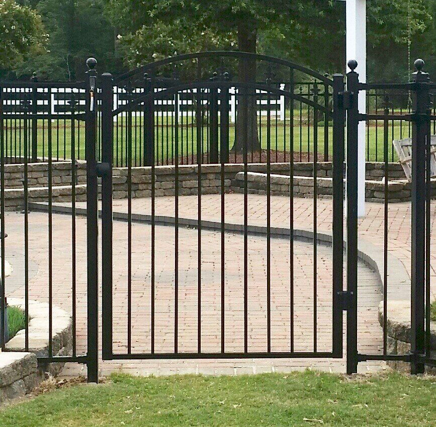 Black metal gate with arched top in a yard with brick pavers and a fence.