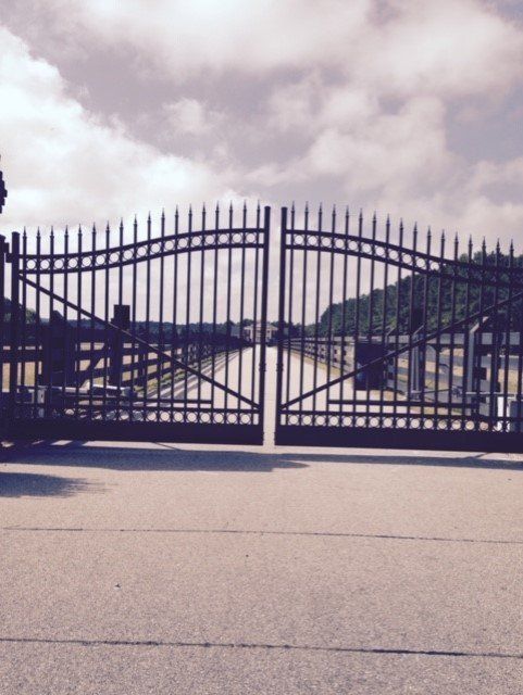 Black wrought iron gates, partially open, lead to a path under a cloudy sky.