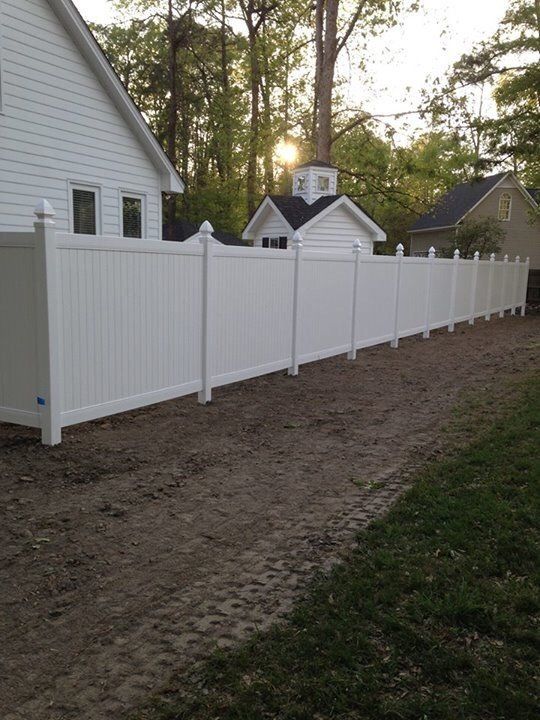 White vinyl fence next to a house and dirt driveway. The sun is shining.