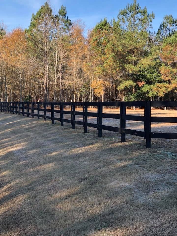 Black wooden fence in a field with brown grass, trees in the background, under a blue sky.