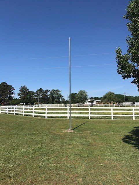 A tall, silver metal pole in a grassy field, white fence and trees in background under a blue sky.