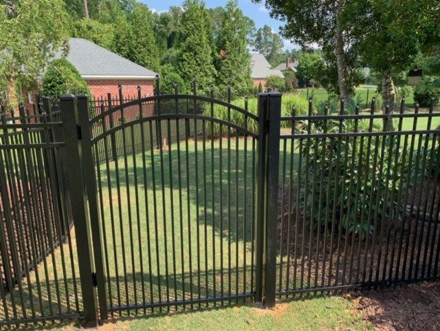 Black metal fence with arched gate in a grassy yard; houses and trees in the background on a sunny day.