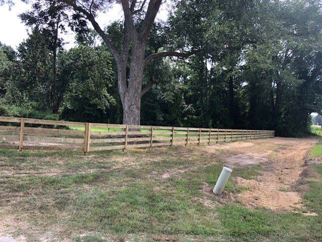 A wooden split-rail fence lines a grassy area next to a road, with trees in the background.
