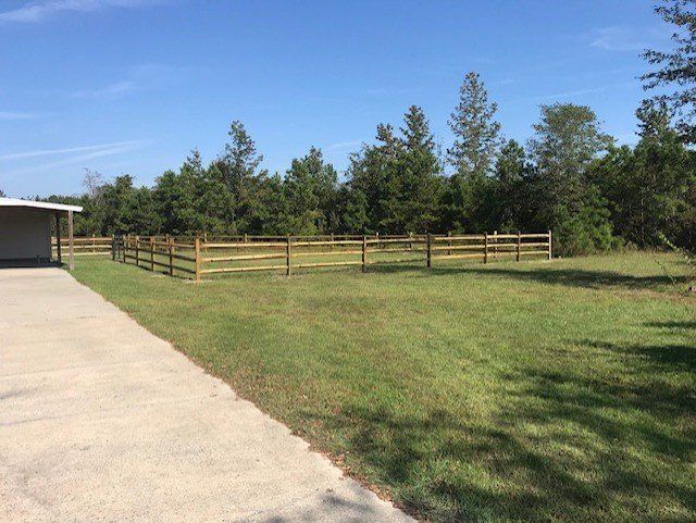 A wooden split-rail fence encloses a grassy area, trees in background, next to a concrete driveway and building.