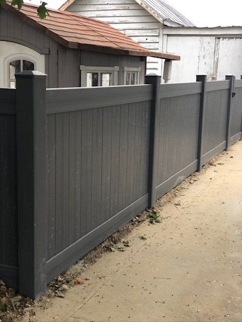 Gray privacy fence in front of a weathered wooden building and a paved surface.