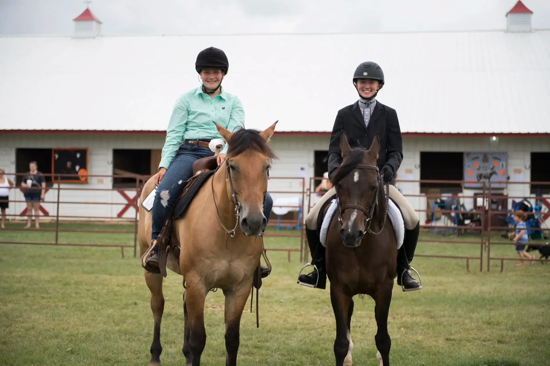 Two riders on horses posing in front of a white barn. One rider wears a green shirt, the other a black coat.