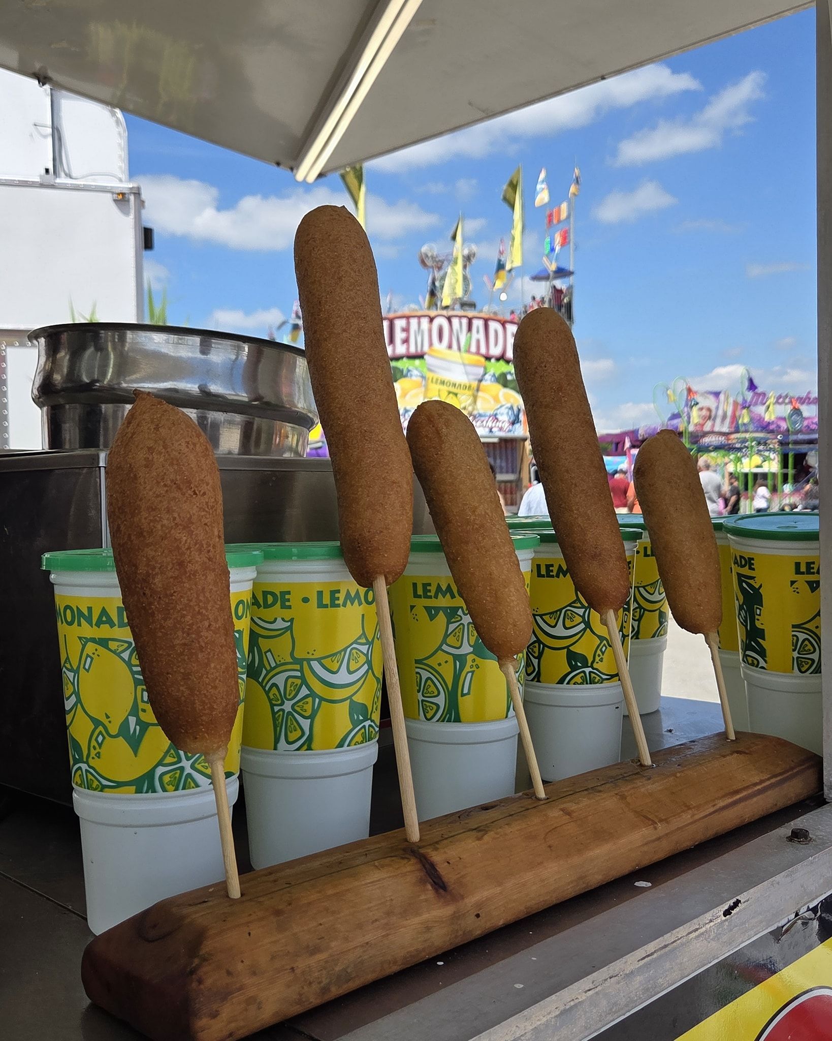 Corn dogs on sticks displayed in front of lemonade cups at a fair. Blue sky backdrop.