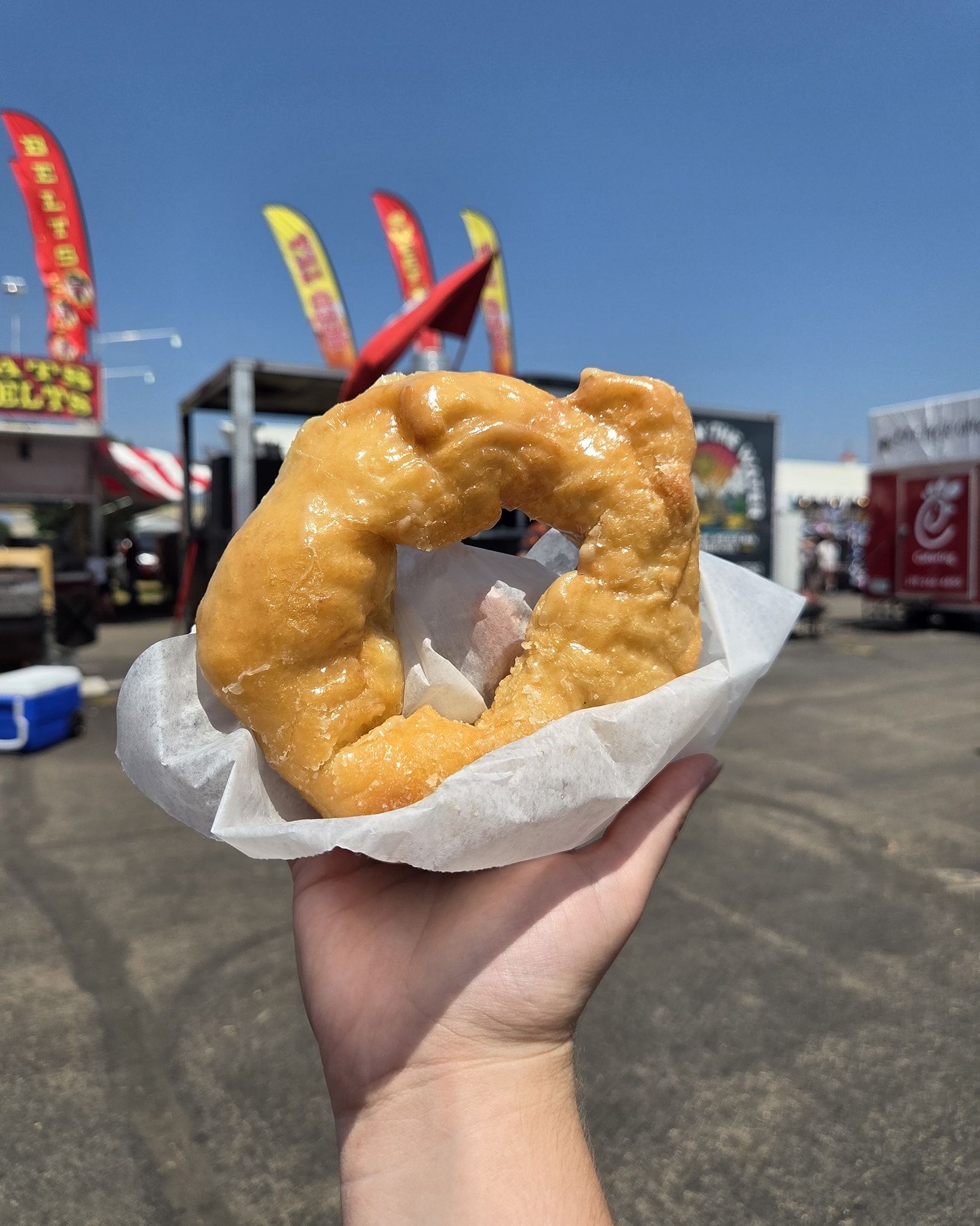 Hand holding a glazed square donut in a paper wrapper at an outdoor fair.