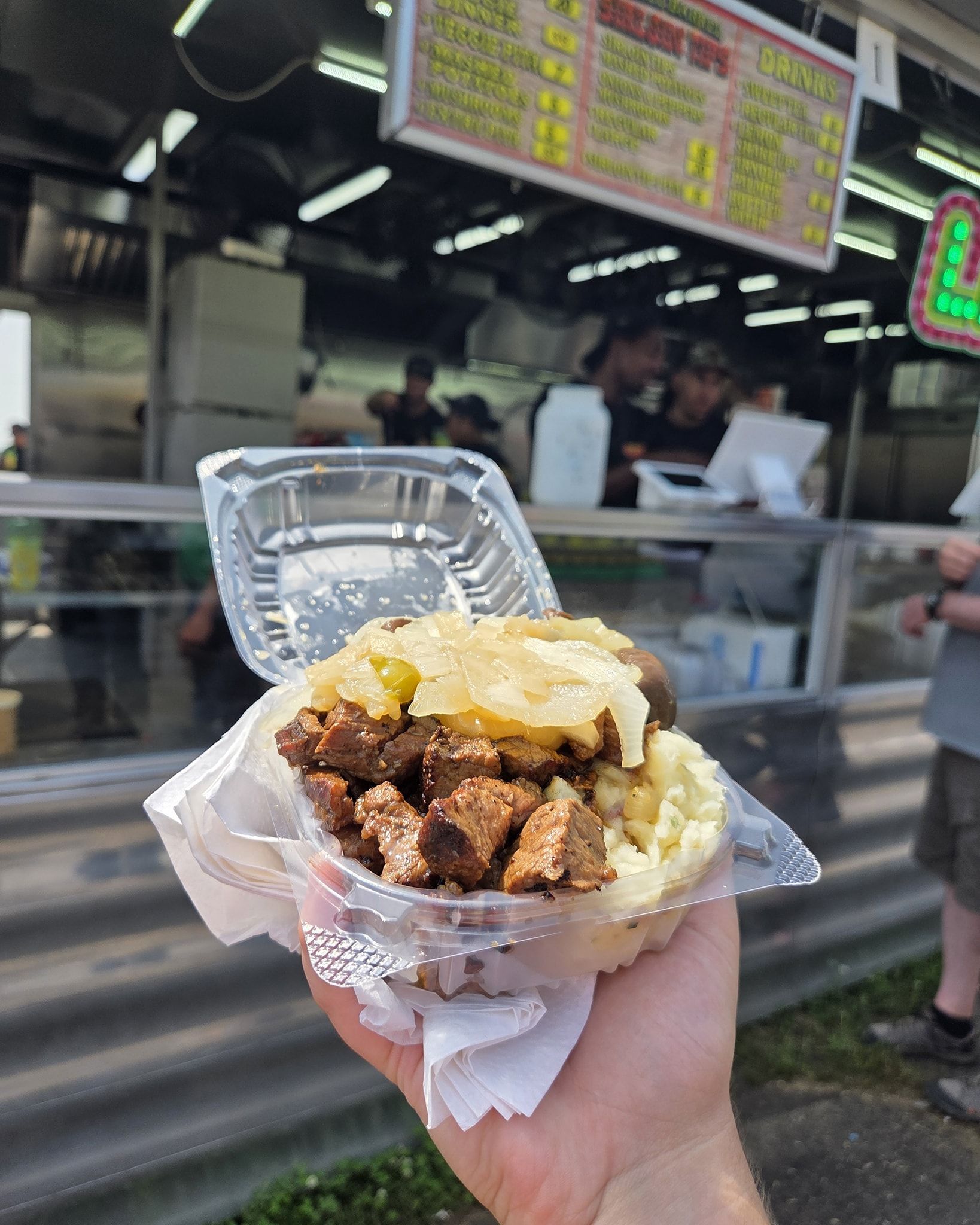 Hand holding a container of food with meat, potatoes, and a topping in front of a food truck.