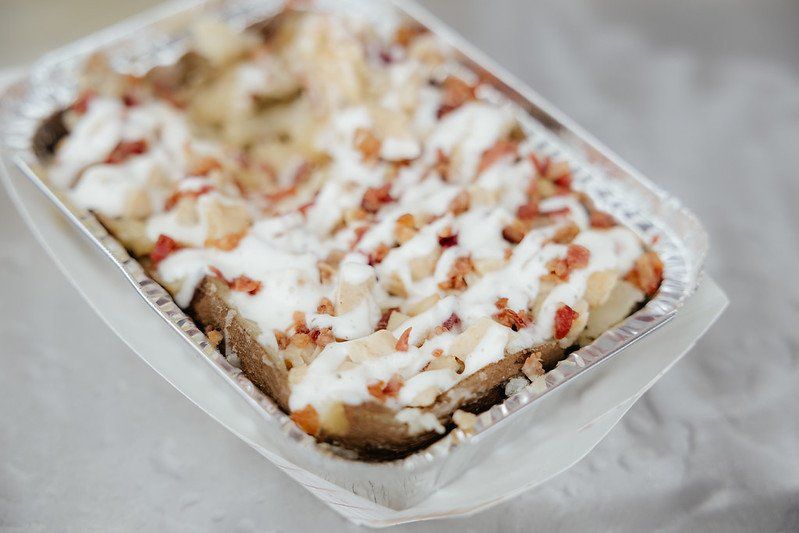 Baked potato in a foil container, topped with bacon and sour cream.