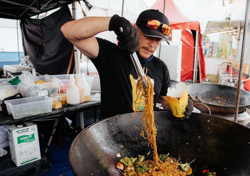 Chef tossing noodles with tongs into a wok. He is wearing a black shirt, cap, and gloves. Outdoor food stall.
