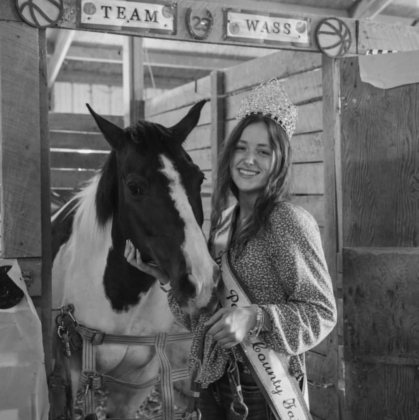 Woman in a crown and sash smiles with a paint horse in a barn stall.