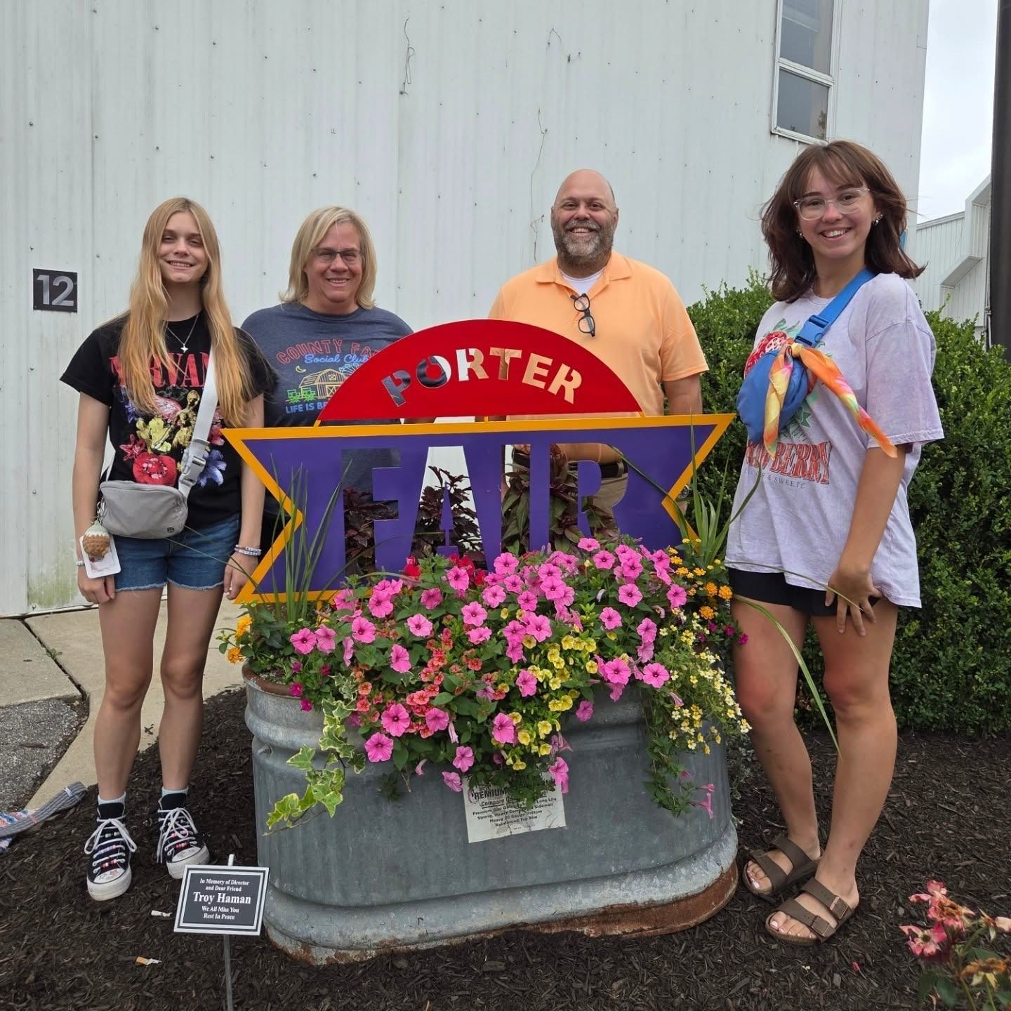 Four people pose in front of a fair sign with flowers. Building in background.