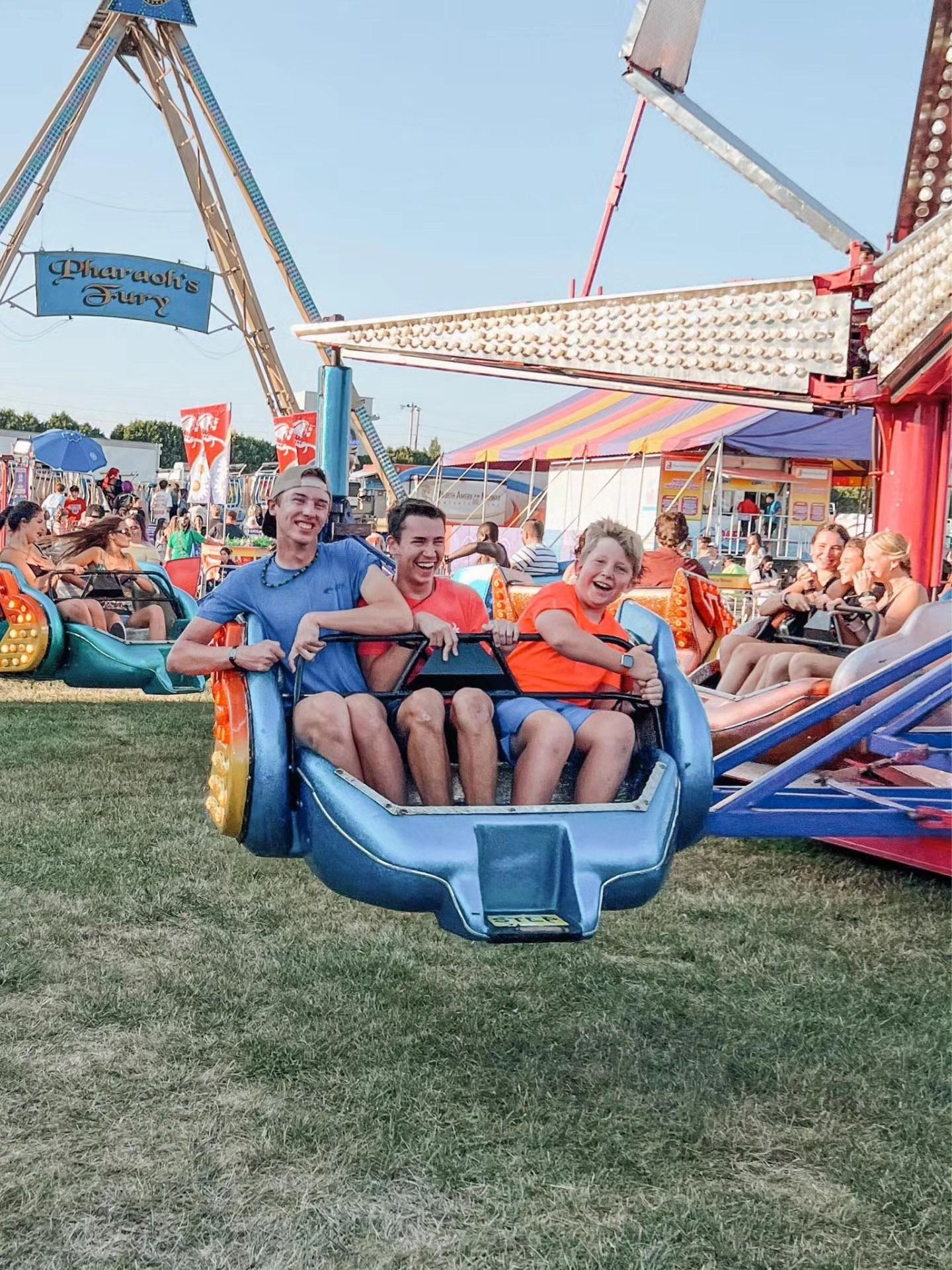 Three people on a carnival ride: blue car, red and orange shirts, big smiles, other rides and people in the background.