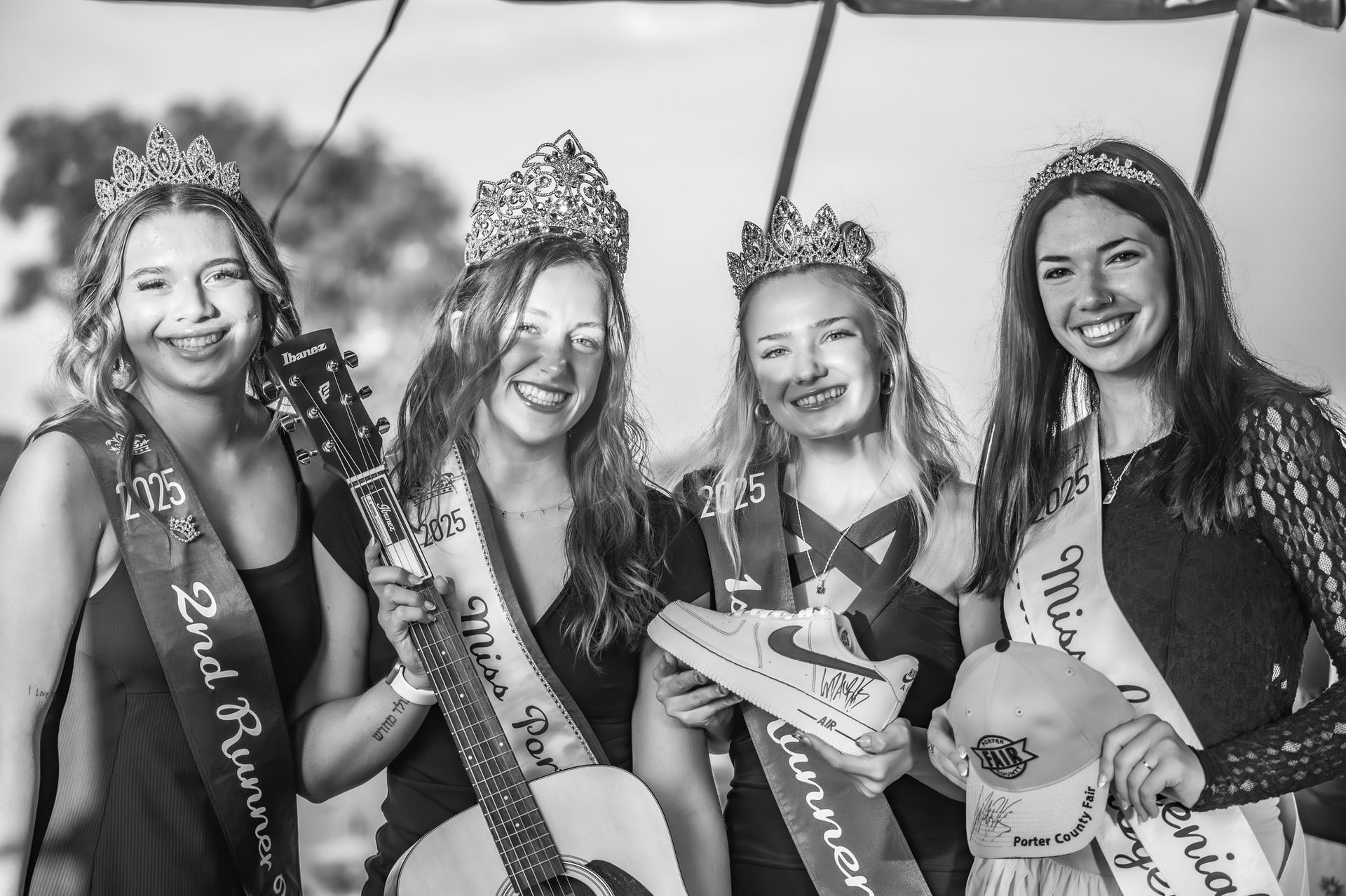 Four women smiling, wearing crowns and sashes, holding a guitar, a shoe, and a hat. Black and white photo.