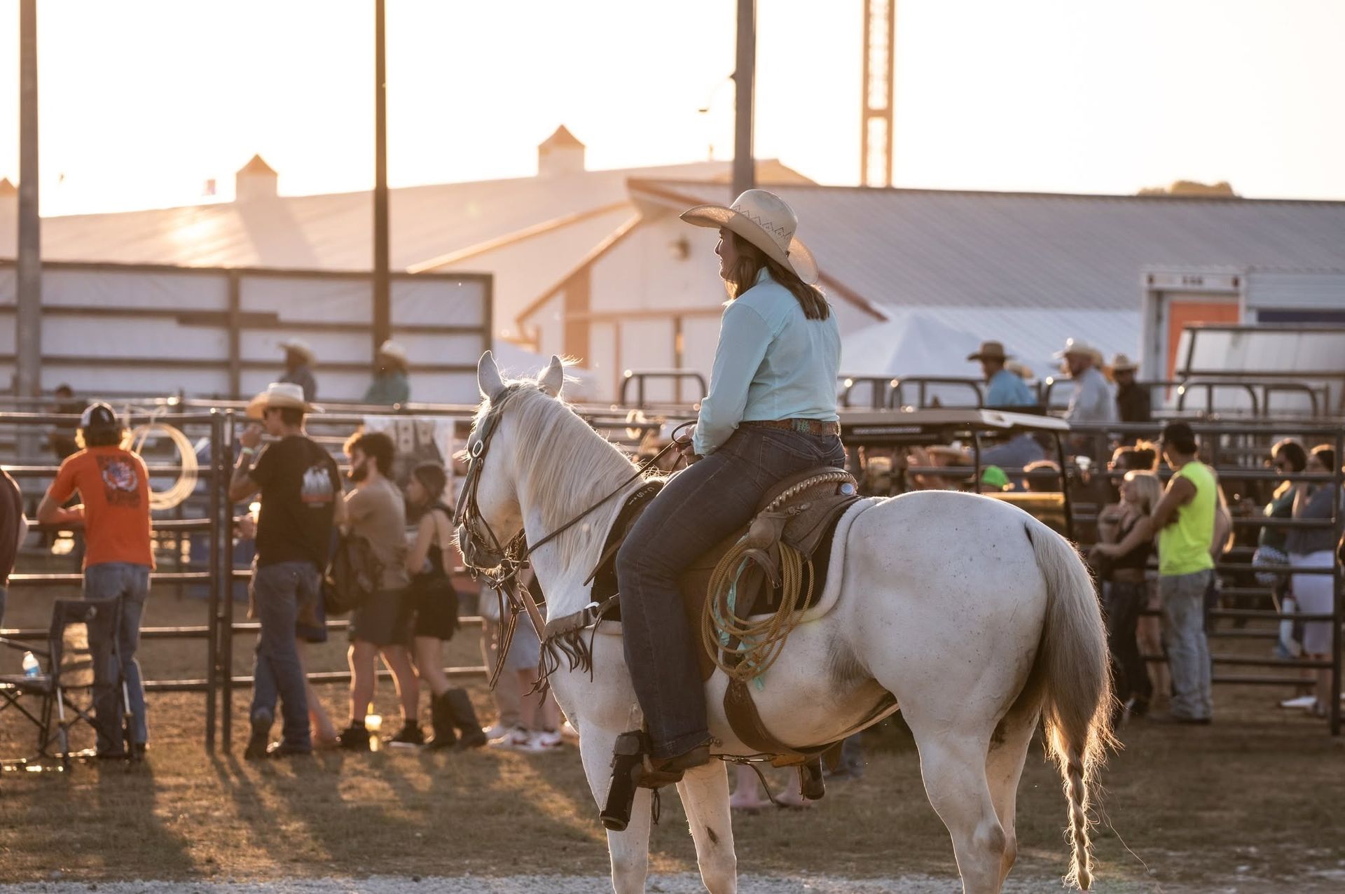 Person on white horse at rodeo, wearing a hat, surrounded by spectators and fences.