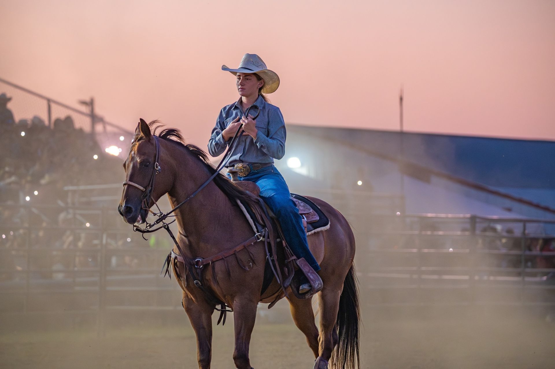 Cowgirl on a brown horse at a rodeo, wearing a cowboy hat and denim, against a dusky sky.