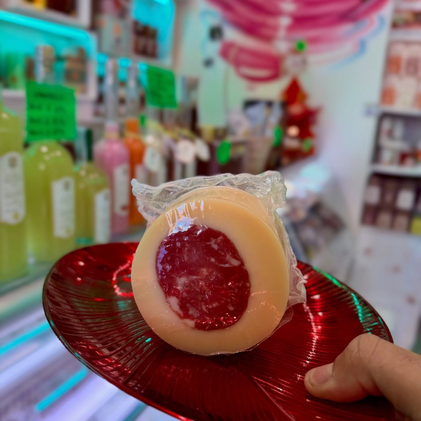 Una persona tiene in mano un piatto rosso con una forma di formaggio avvolta in pellicola trasparente e al centro scuro, con delle bottiglie sullo sfondo.