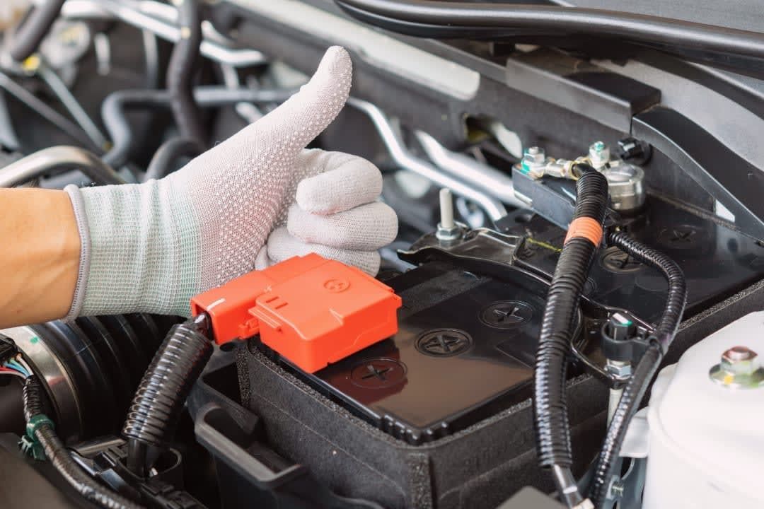Gloved hand giving thumbs up near car battery, indicating it's working properly. Orange cover on positive terminal.