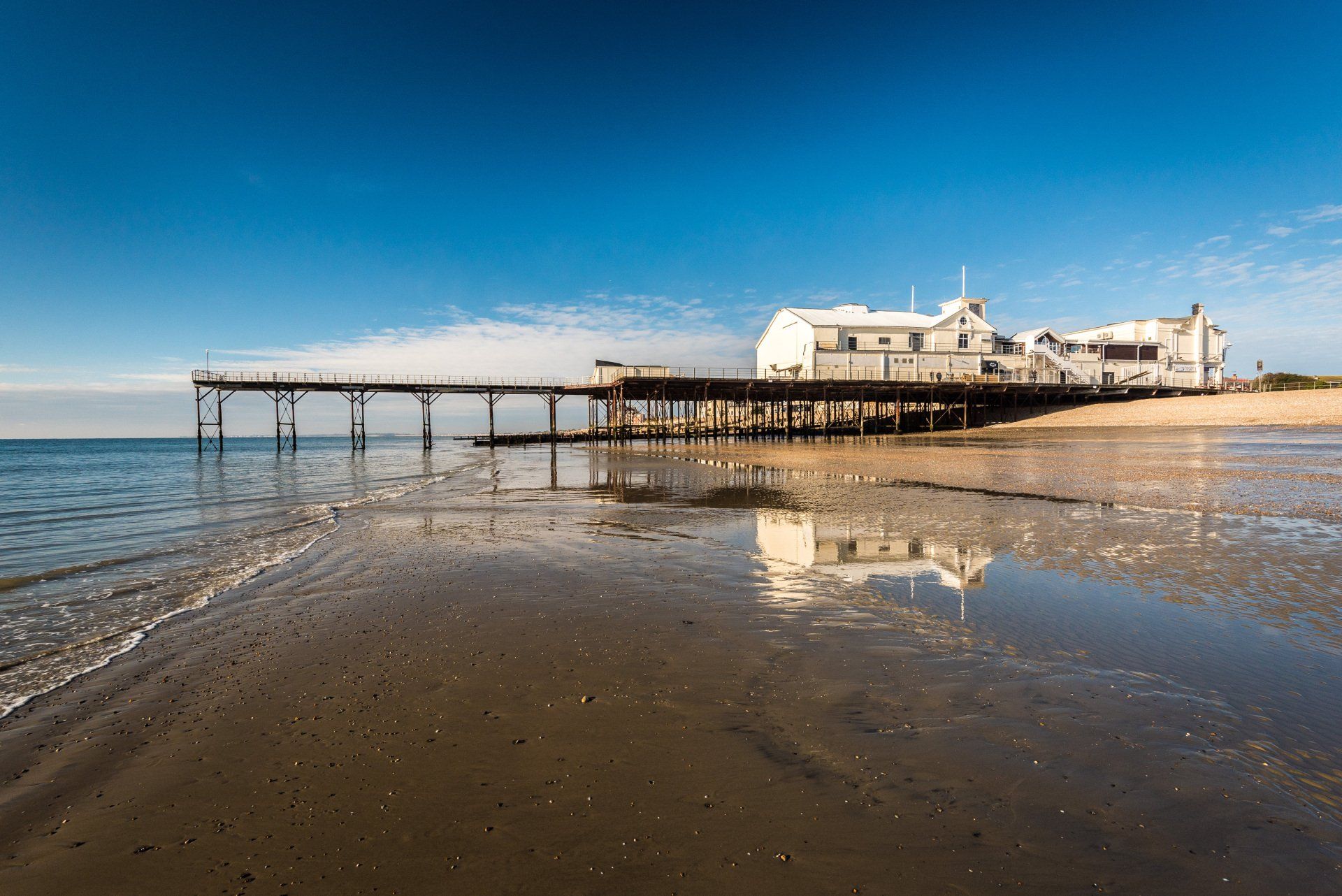 Bognor Regis Pier, reflected in the wet sand at low tide and blue sky beyond.