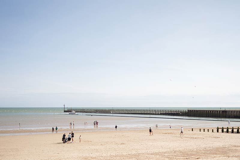 A view across the shingle beach to low tide at one of the less busy beaches in Littlehampton, with just a few people enjoying the beach and water.