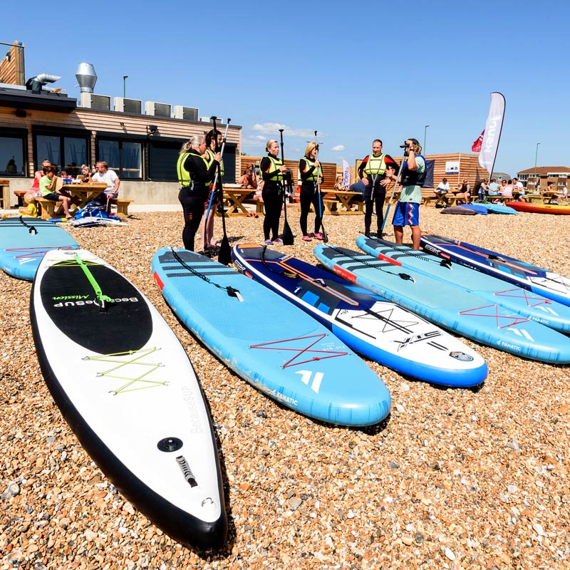 Water Sports students with their instructor and paddleboards on the shingle beach by the The Beach @Littlehampton Activity Hub and Cafe.