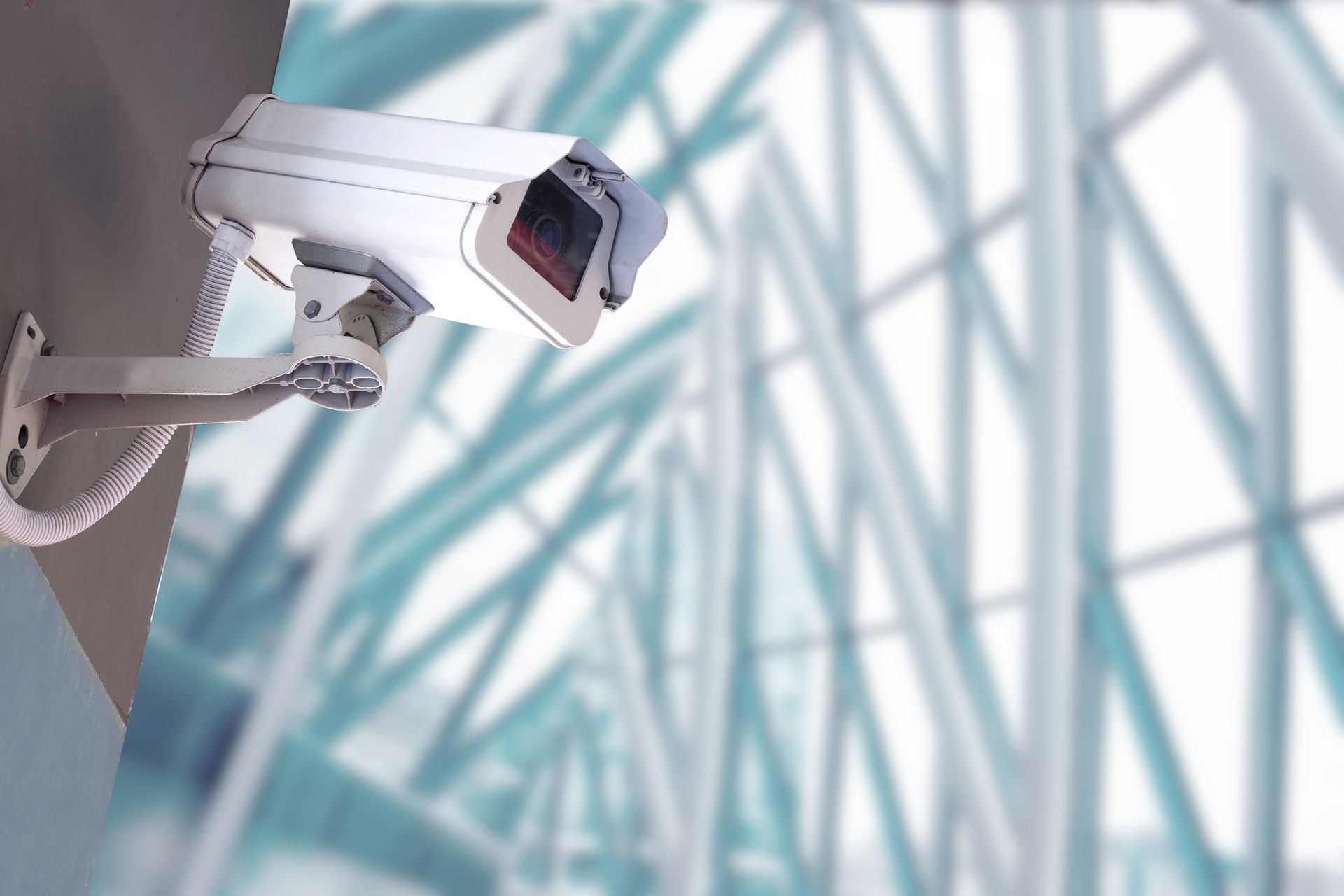 Wall-mounted security camera with conduit near modern glass beams.