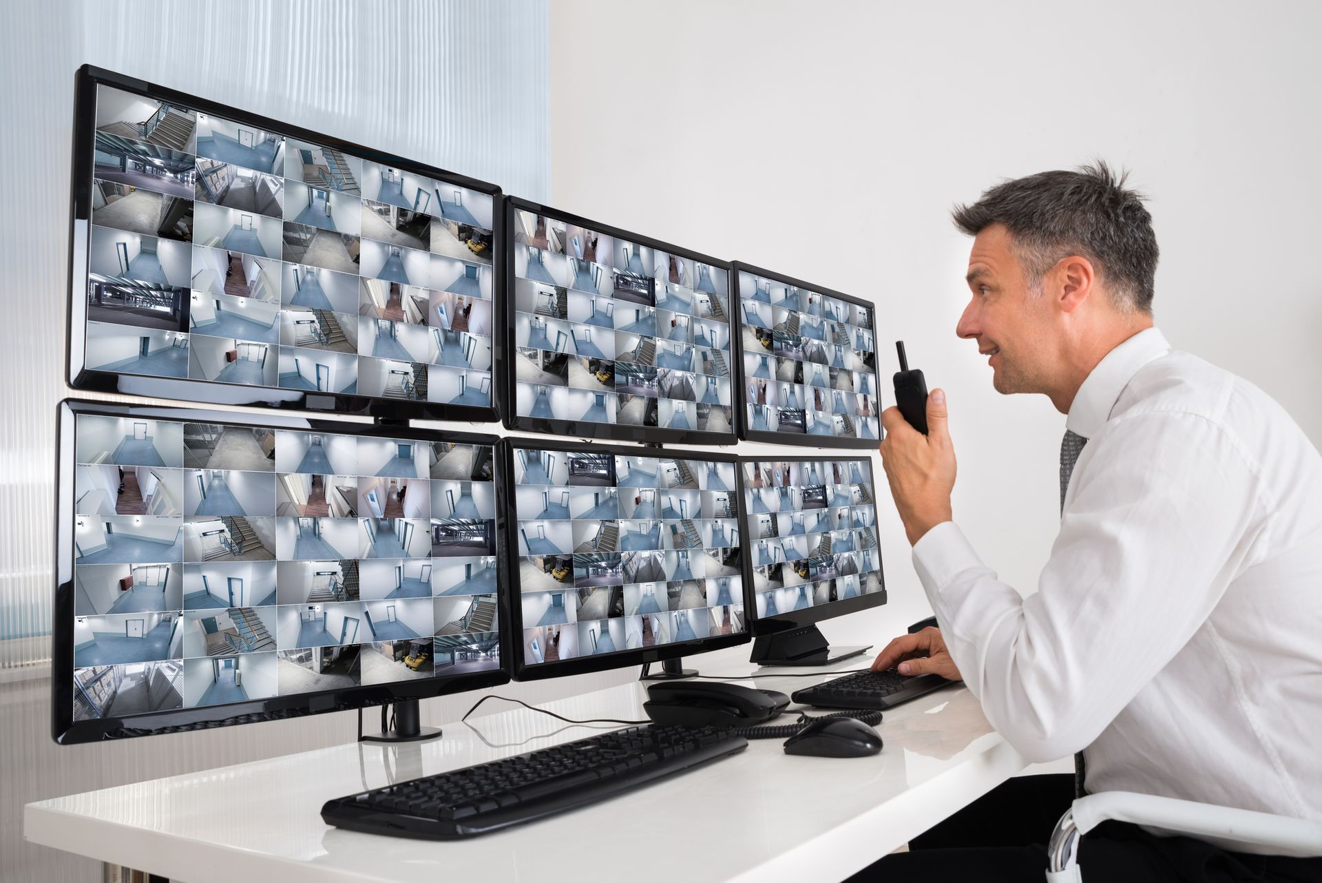 A man is sitting at a desk looking at a bunch of monitors and talking on a walkie talkie.