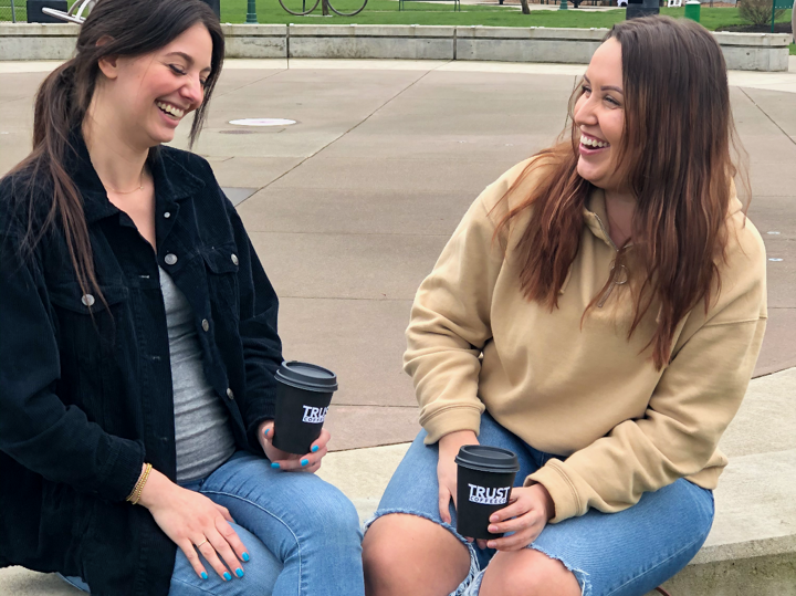 Two women are sitting next to each other holding cups of trust coffee and laughing