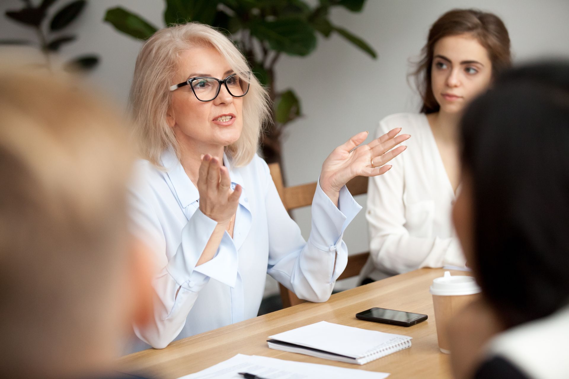 Woman with glasses gestures while speaking at a table, people listen.