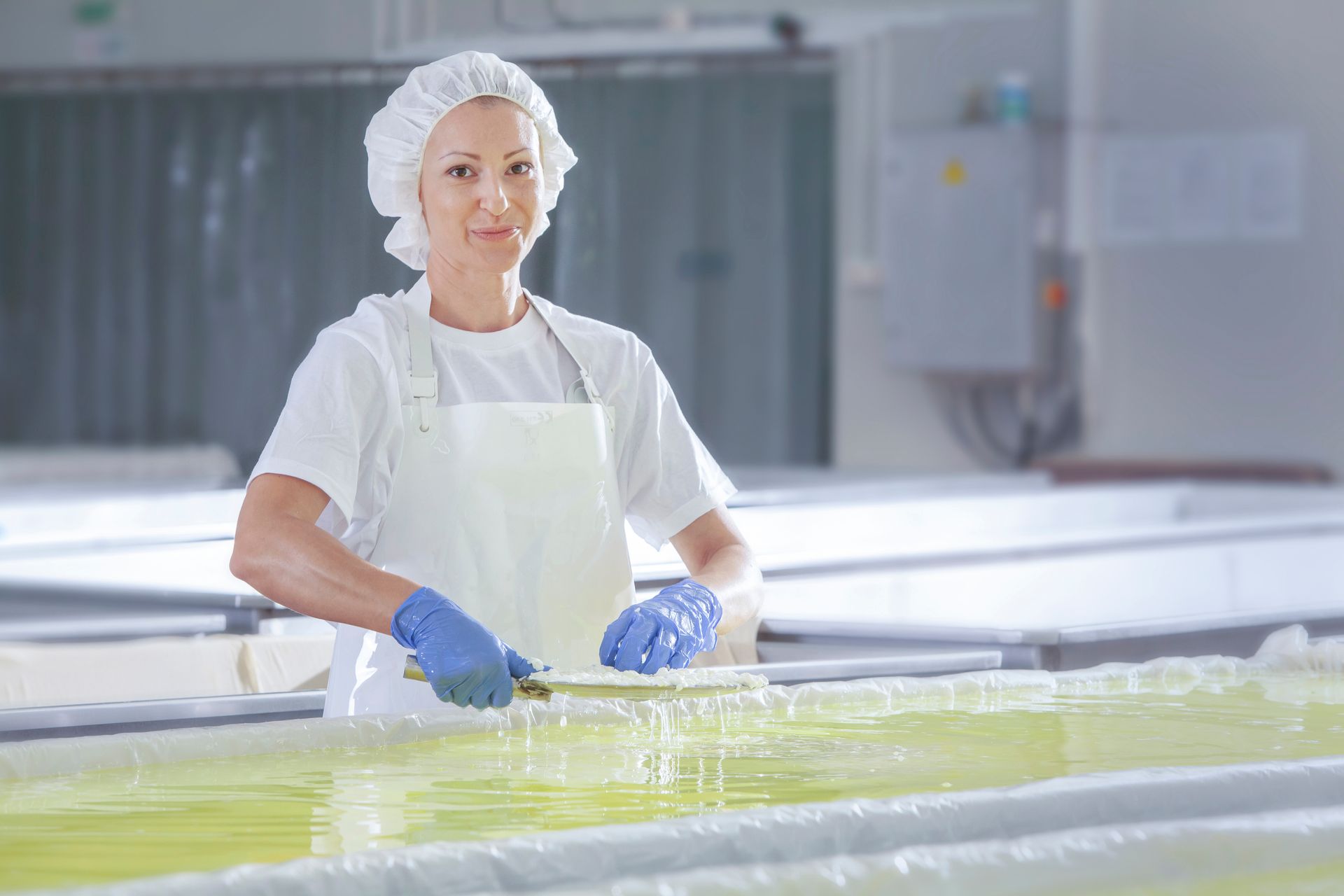 Woman in white uniform and hair net, blue gloves, processing food in a facility, smiling.