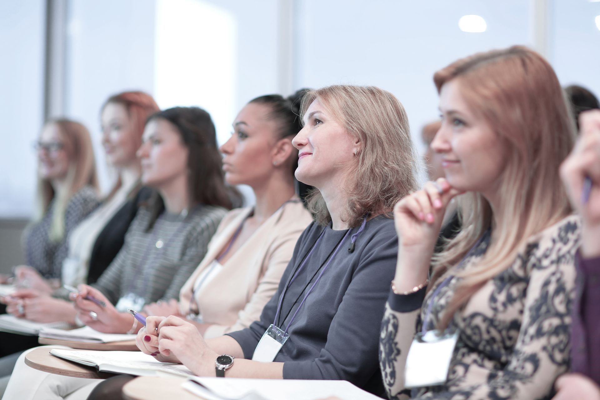 Attendees at a seminar listen intently, some with pens, in a well-lit room.