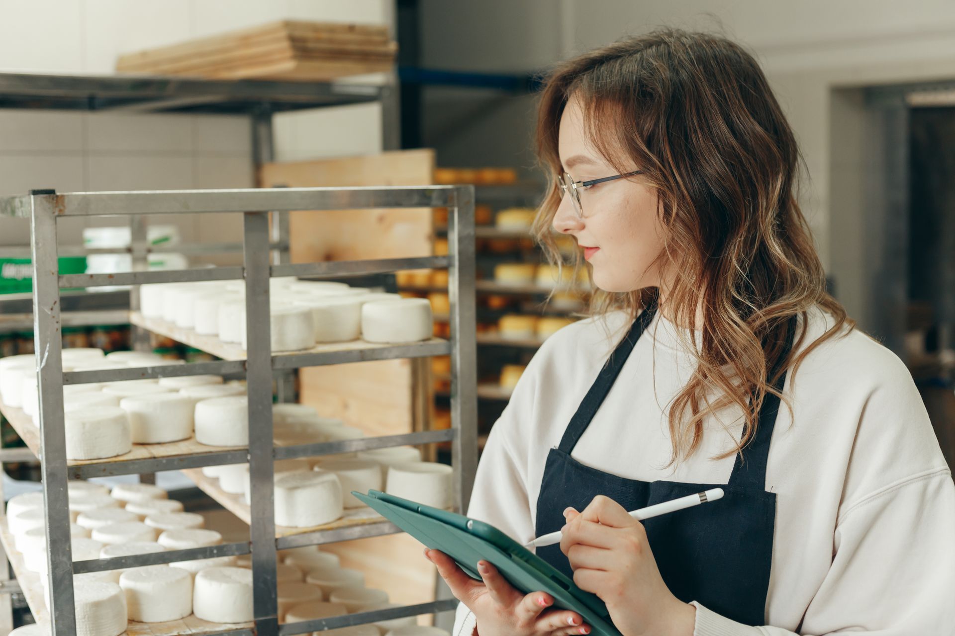 Woman in apron and glasses, examining cheese on shelves with a tablet and stylus.