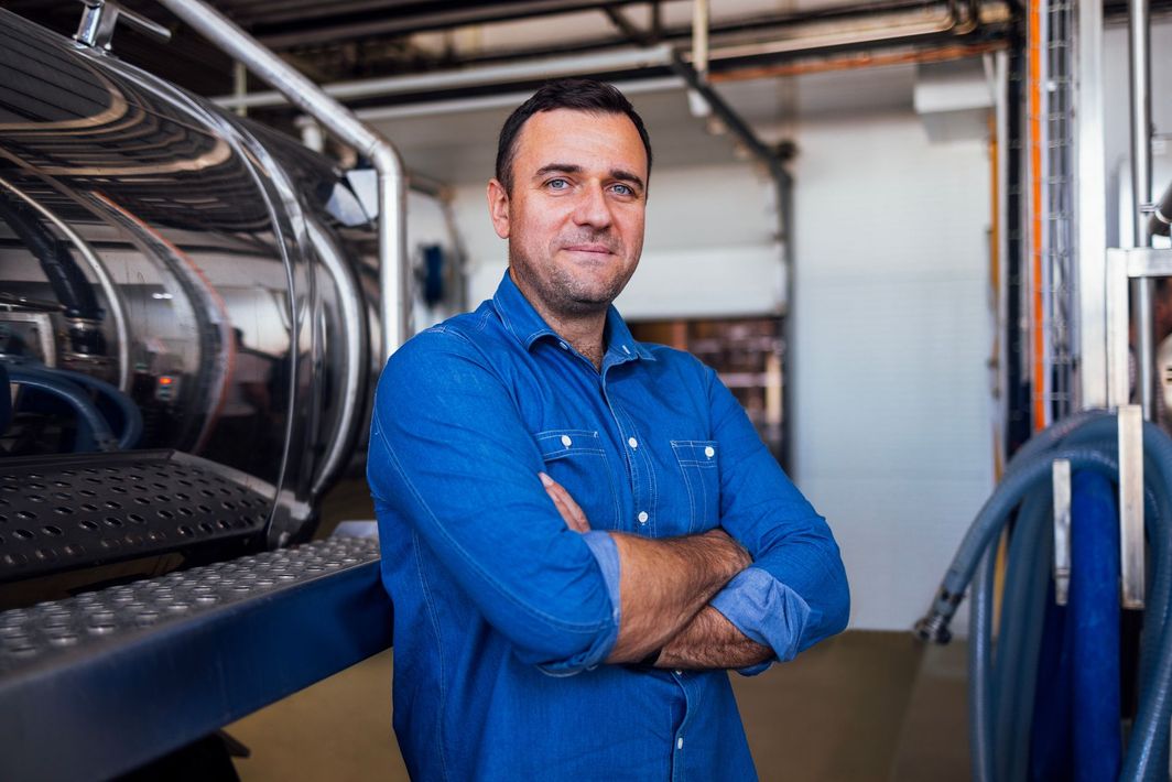 Man in blue shirt, arms crossed, stands in front of brewery equipment.