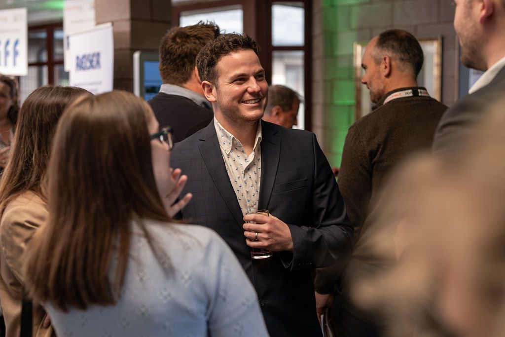 Crowd of people in an event space, blurred. Booths and signs in the background.