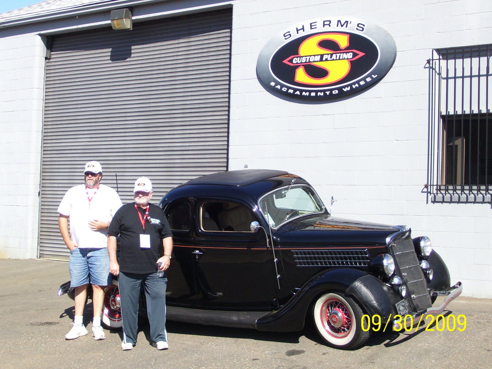Two men standing next to a black car in front of a building that says sherms