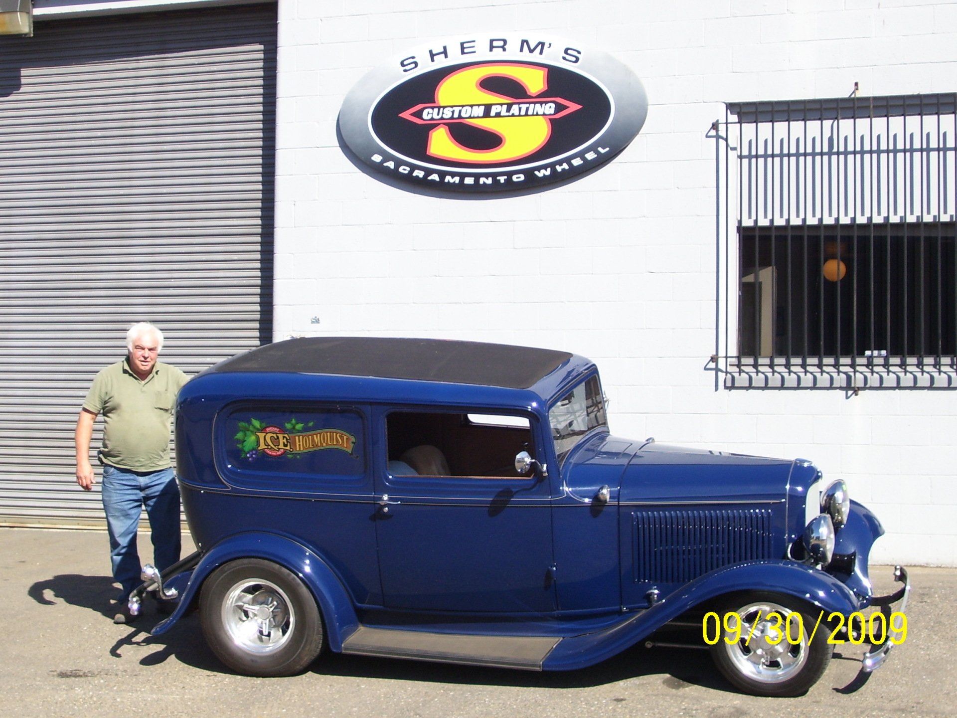 A man stands next to a blue car in front of a sign that says sherman 's